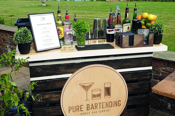 Outdoor mobile bar set up with bottles of alcohol on top and a wooden circular logo sign for Pure Bartending